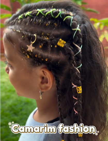 Criança com penteado de tranças decoradas com fitas coloridas, glitter dourado e enfeites de borboleta e estrela